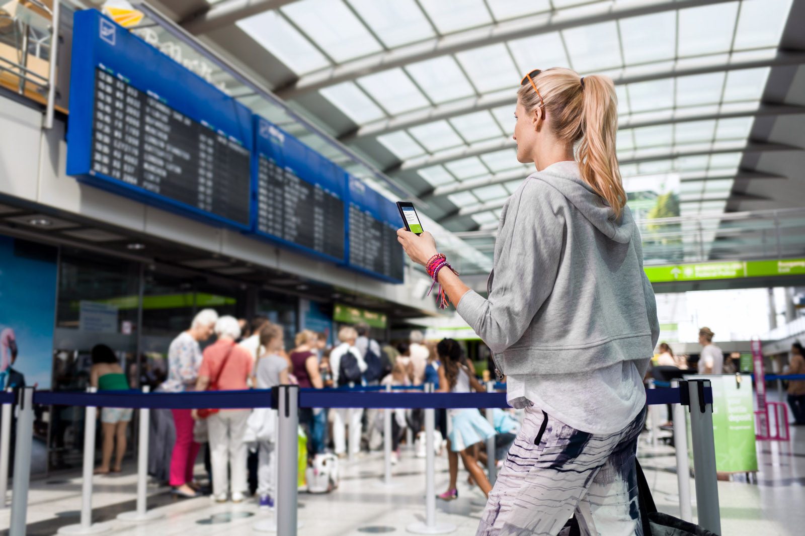 Frau steht am Flughafen, schaut auf ihr Smartphone und die Anzeigetafel